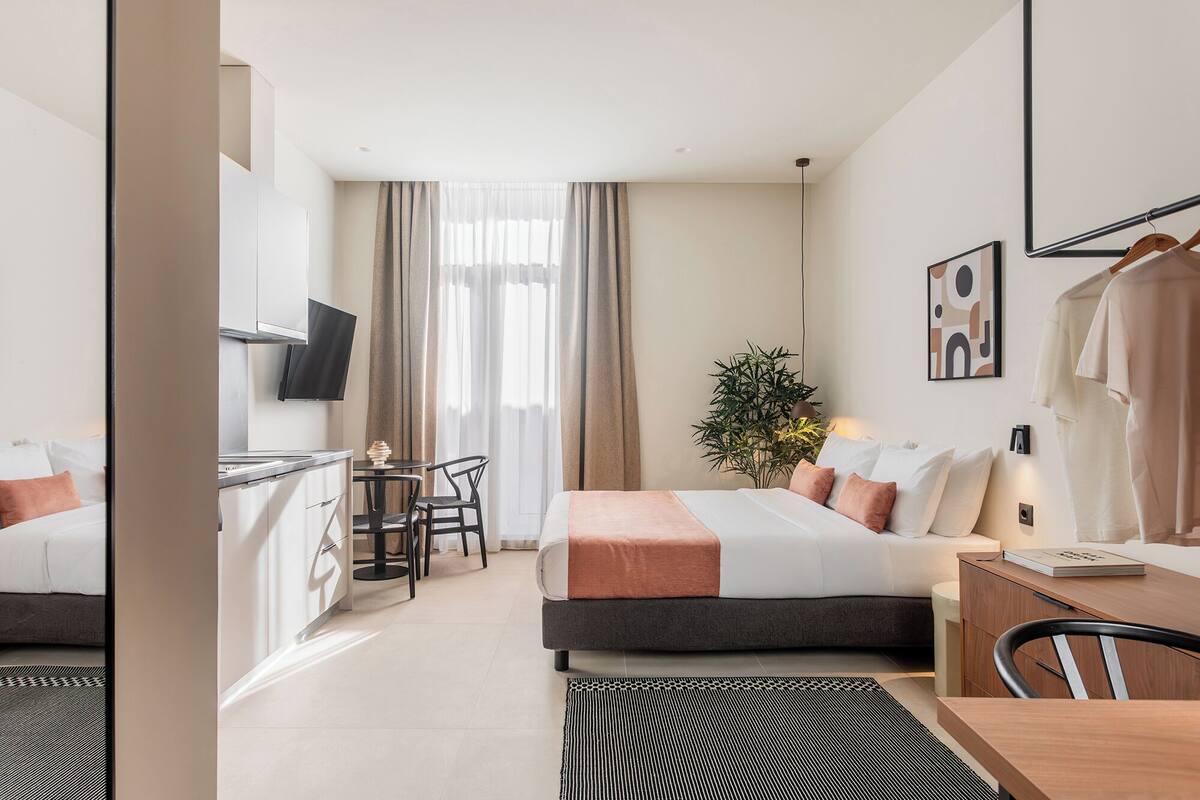 A modern hotel room with a bed, bedside table, desk, chair, and a view of the city.