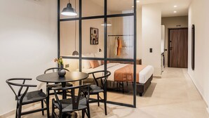A modern hotel room with a glass partition, a round dining table with chairs, a bed with a white and orange bedspread, and a visible bathroom with a towel rack.