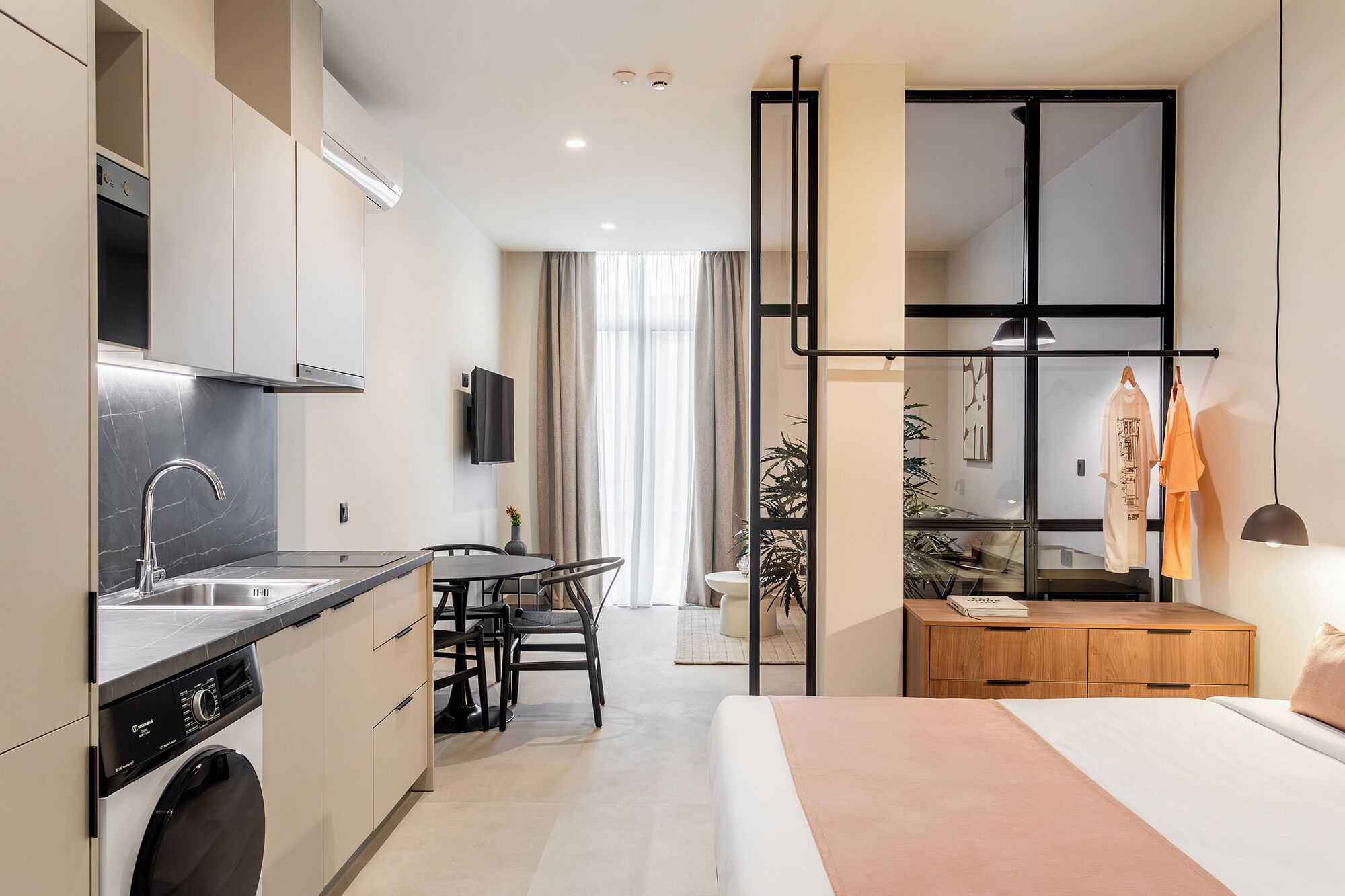 A modern hotel room with a kitchen, a dining area, a large window, and a bed.