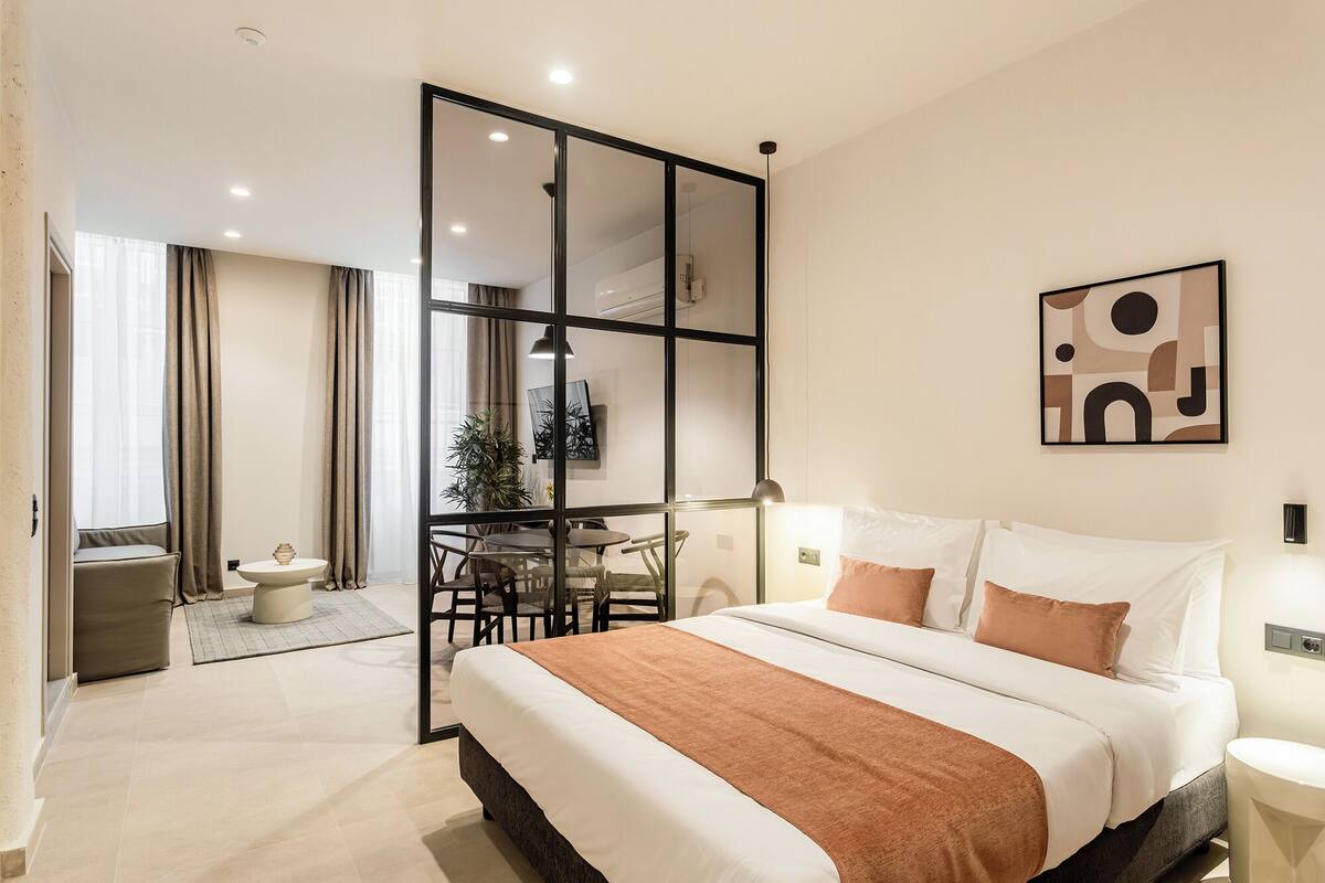 A modern hotel room with a large bed, a dining area, and a sliding glass door leading to a balcony.