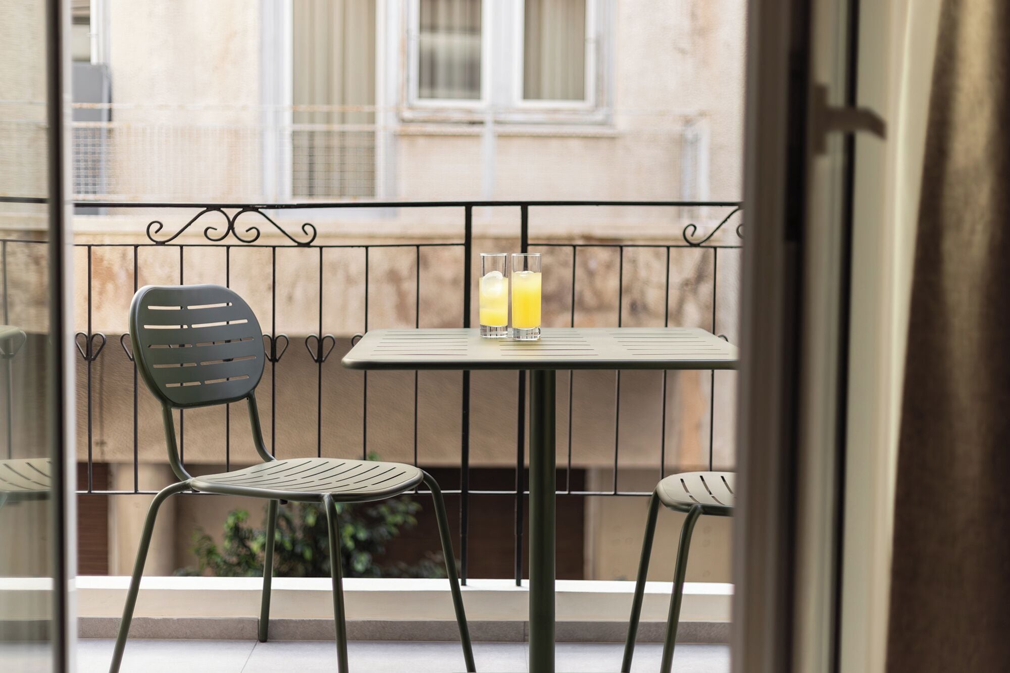 A balcony with a table and two chairs, two glasses of orange juice, and a view of a building.