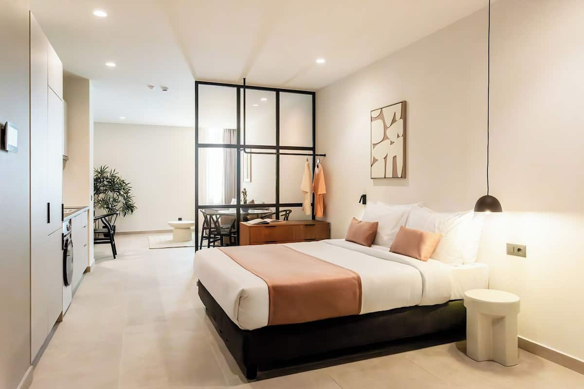 A modern bedroom with a large bed, bedside table, and a small stool. There is a desk with chairs and a plant in the adjacent room.
