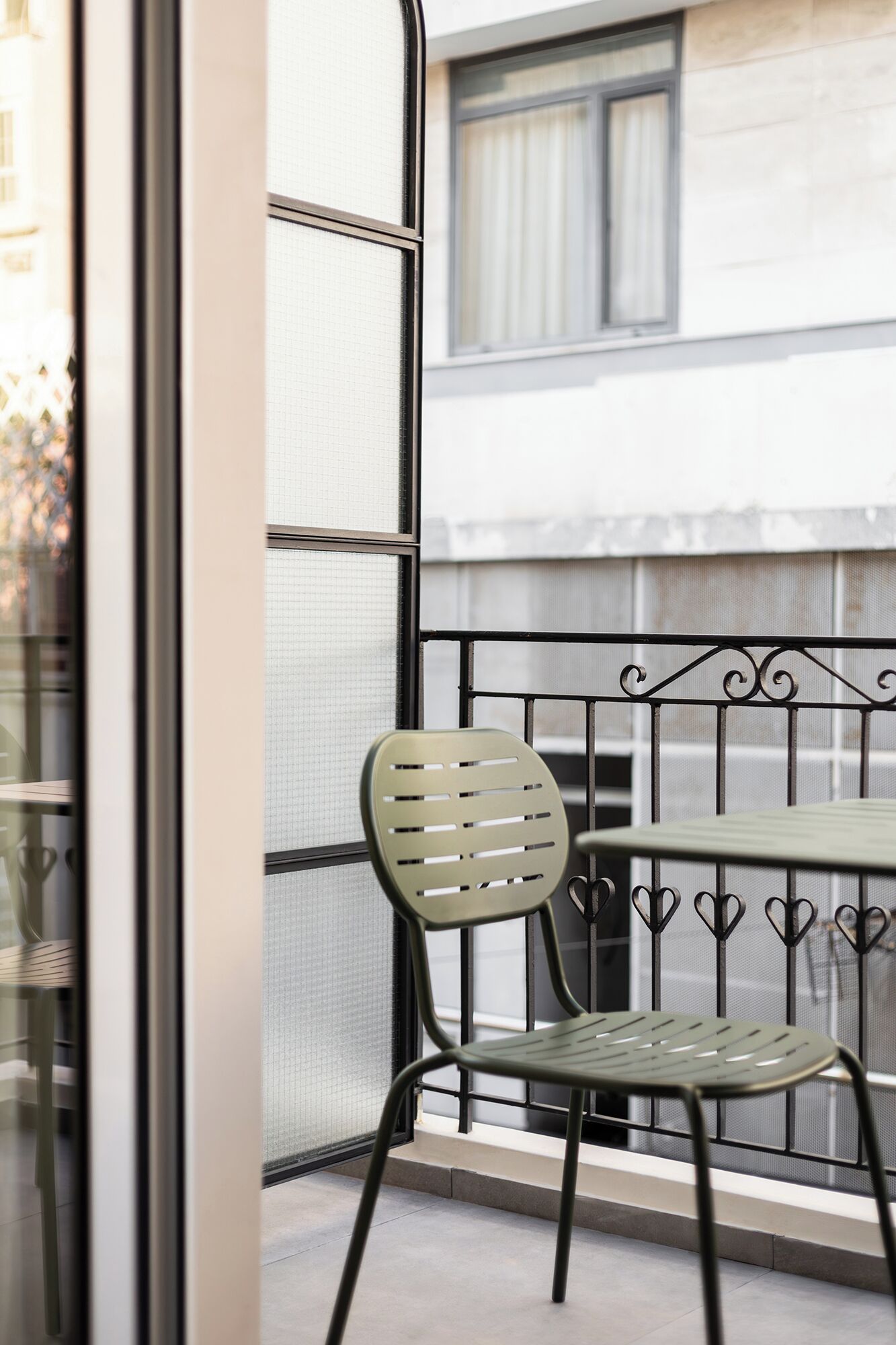 Un balcón con una silla y una mesa de metal, una ventana con vistas a edificios y una barandilla de metal.