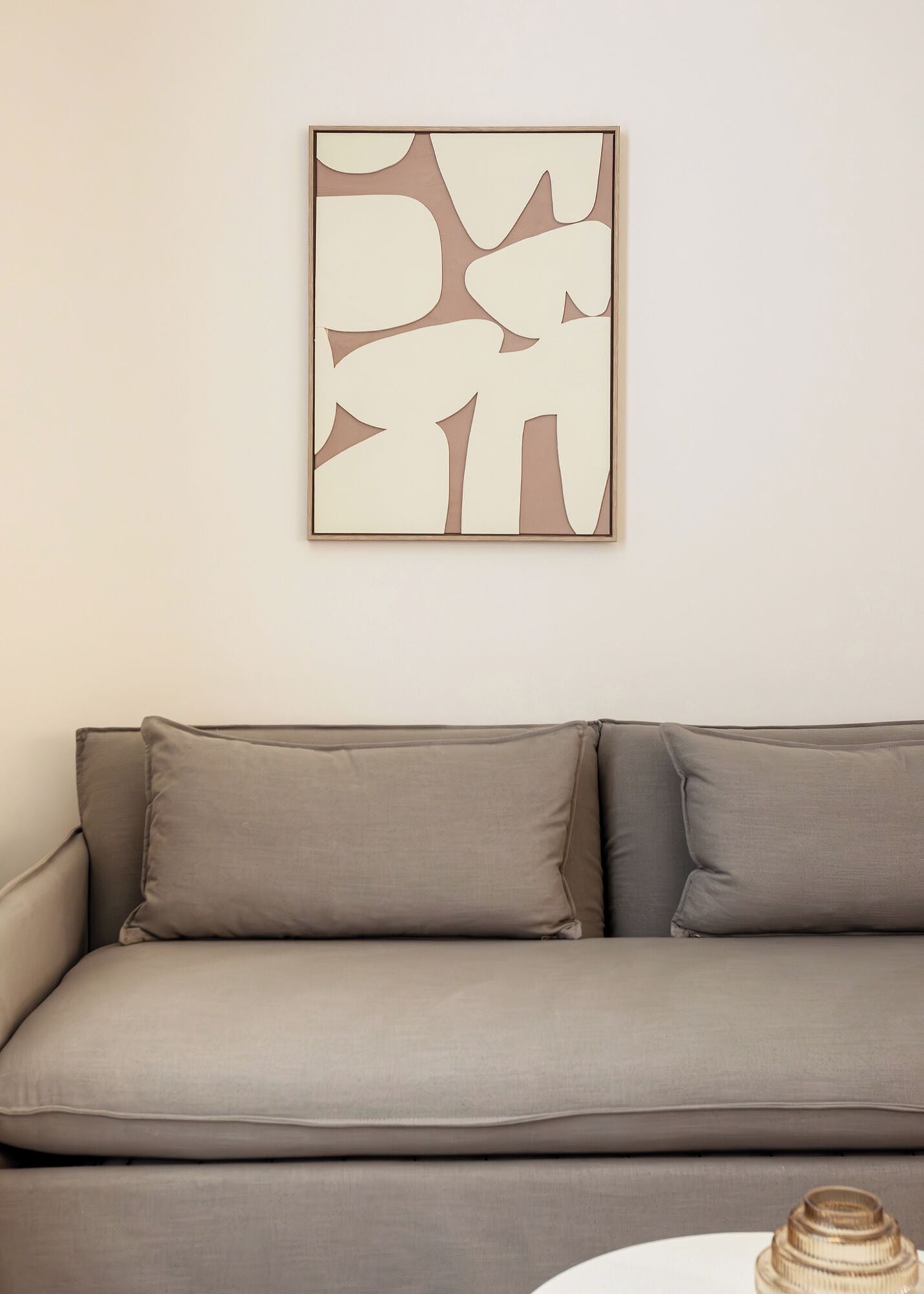 Un sofá de tonos neutros con un diseño sencillo y moderno, ubicado frente a una pared lisa con una obra de arte abstracta enmarcada.