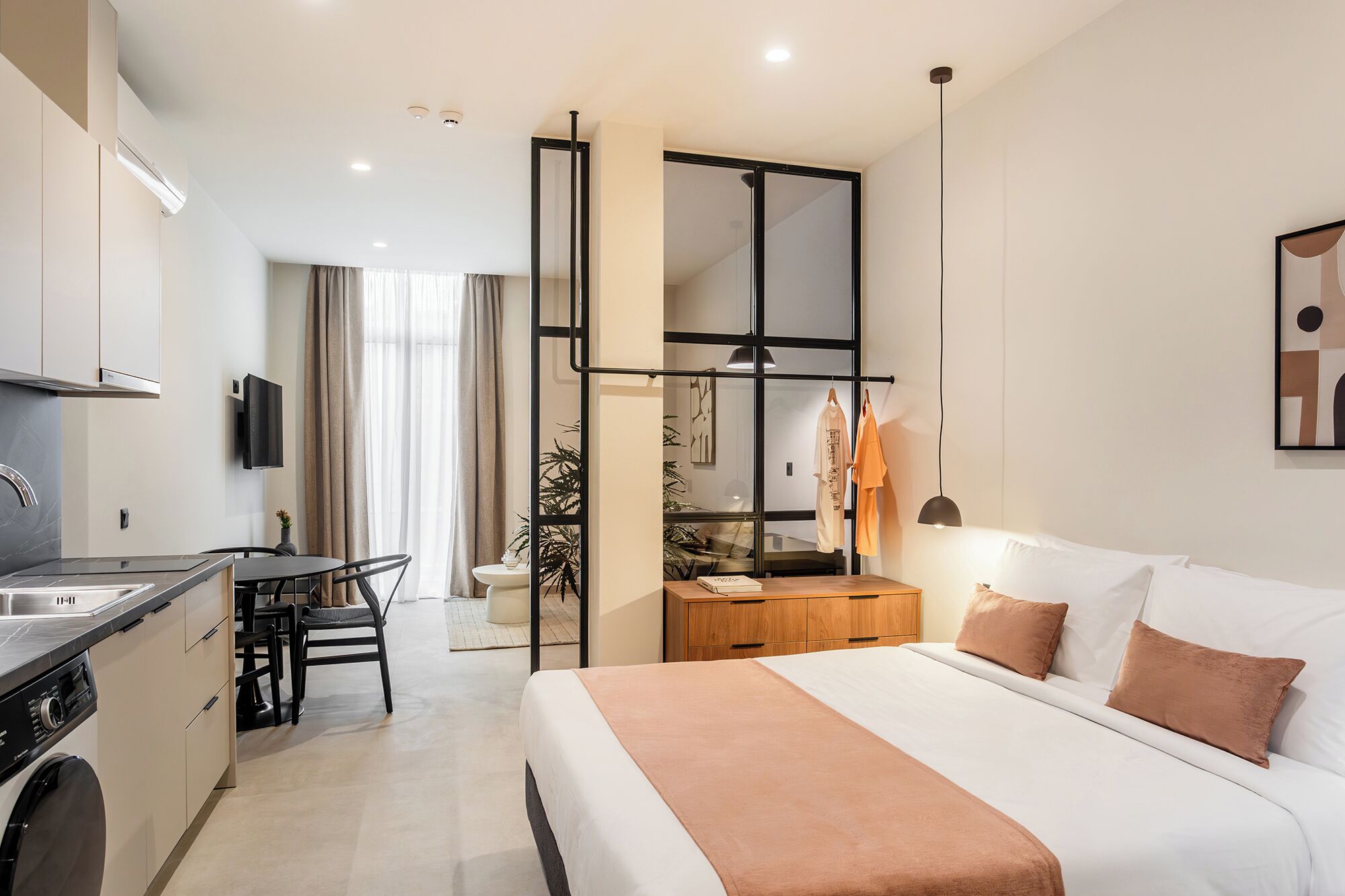 A modern hotel room with a bed, a desk, a chair, a small kitchen area, and a large window.