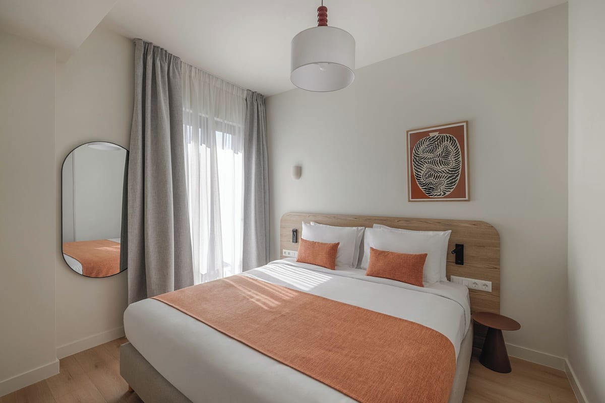 A modern bedroom with a large bed, a bedside table, a round mirror, and a wall-mounted light fixture.