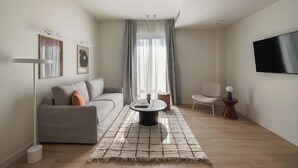 Ein modernes Wohnzimmer mit einer Couch, einem Couchtisch, einem an der Wand montierten Fernseher und einem großen Fenster mit Vorhängen.
