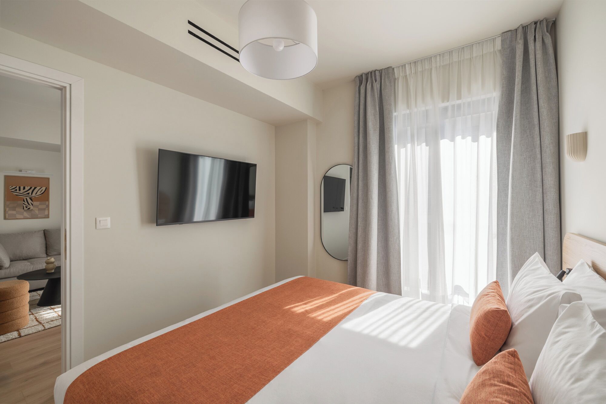 A modern hotel room with a bed, a flat-screen TV, a mirror, and a window with curtains.