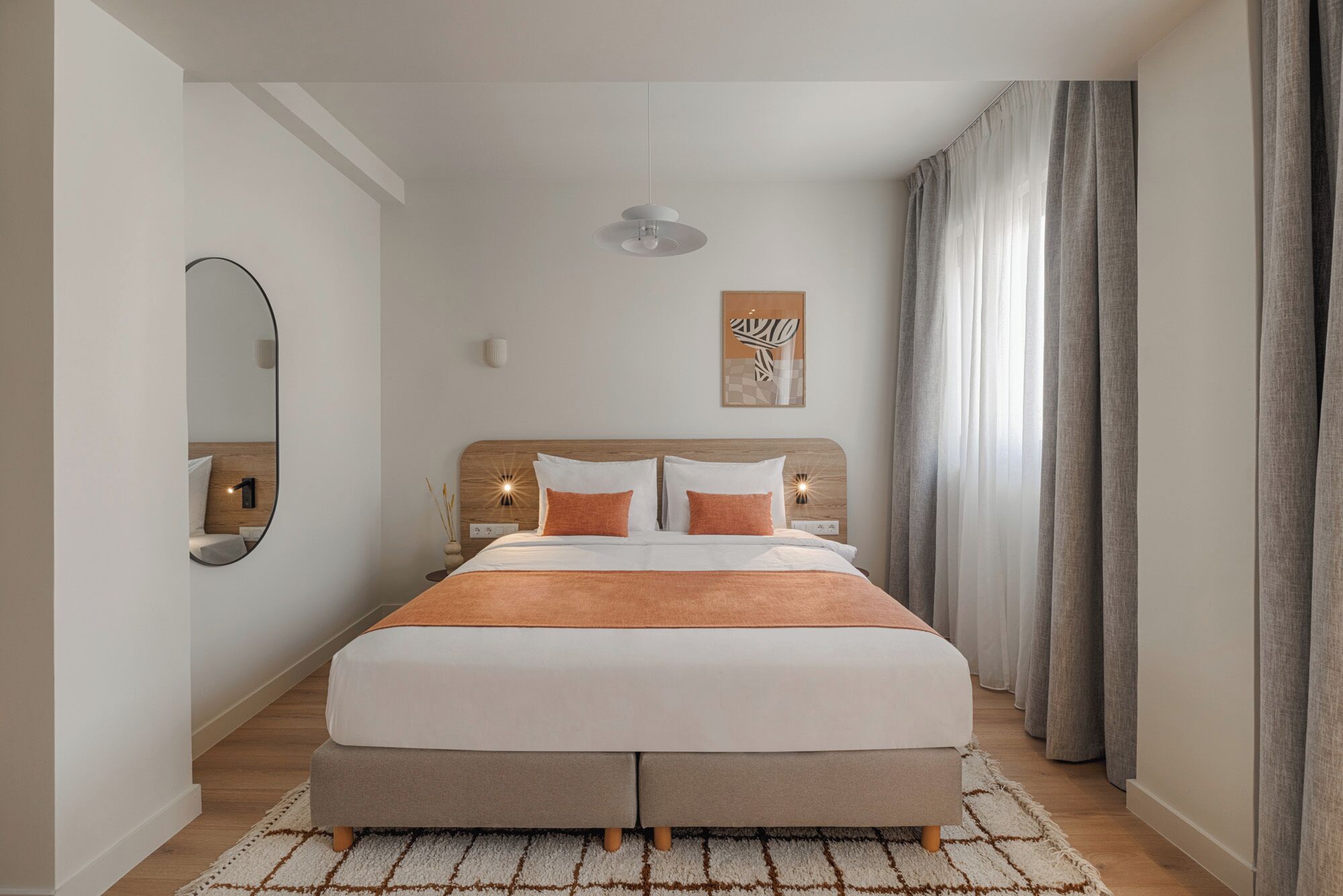 A bedroom with a bed, a bedside table, a wall-mounted light, a mirror, and a window with curtains.