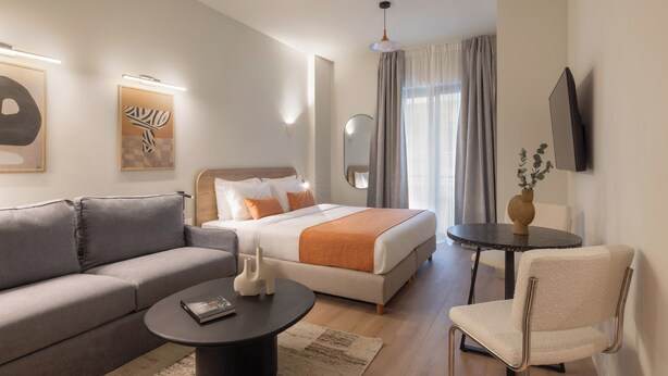 Ein modernes Hotelzimmer mit Bett, Sofa, Sessel, rundem Couchtisch und einem an der Wand montierten Fernseher.