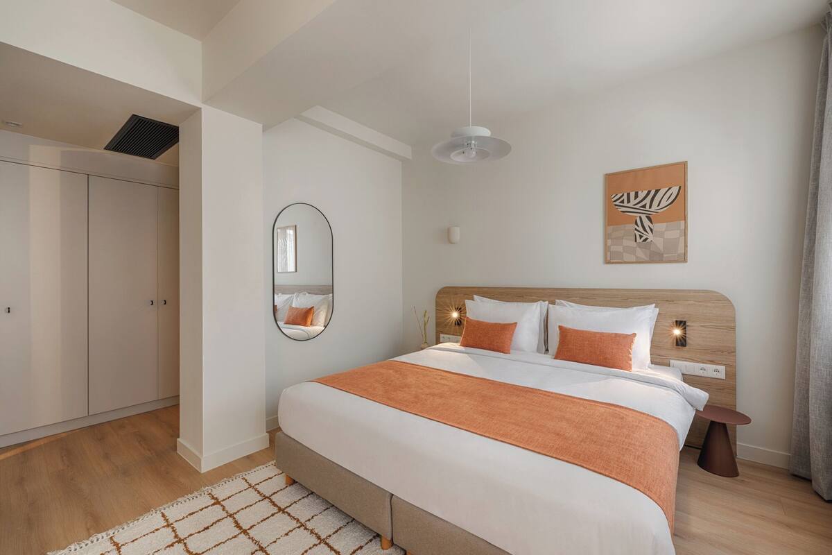 A bedroom with a bed, a mirror, a bedside table, a nightstand, a wall-mounted light, and a ceiling vent.