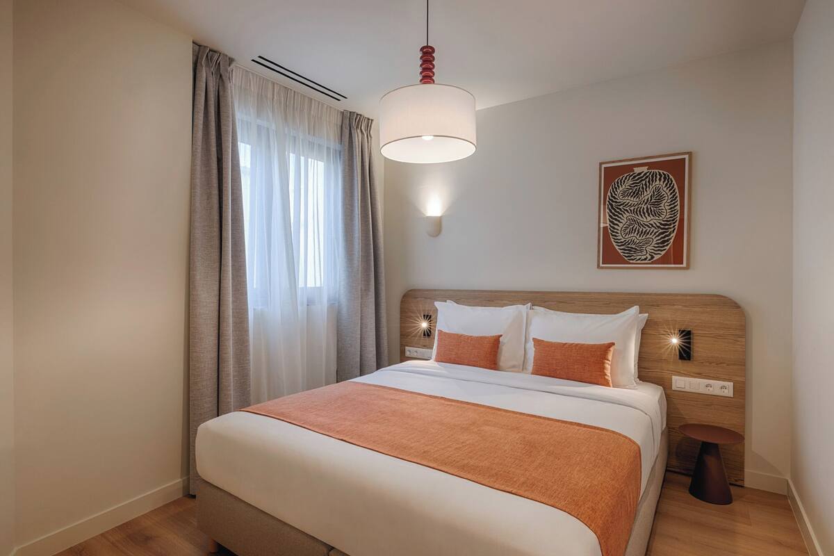 A bedroom with a bed, curtains, a wall-mounted light, a decorative piece, and a bedside table.