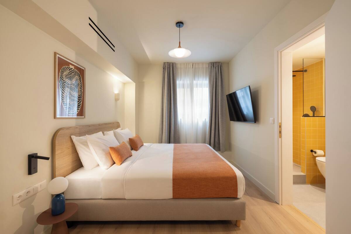 A modern hotel room with a bed, a flat-screen TV, a bathroom with yellow tiles, and a painting on the wall.