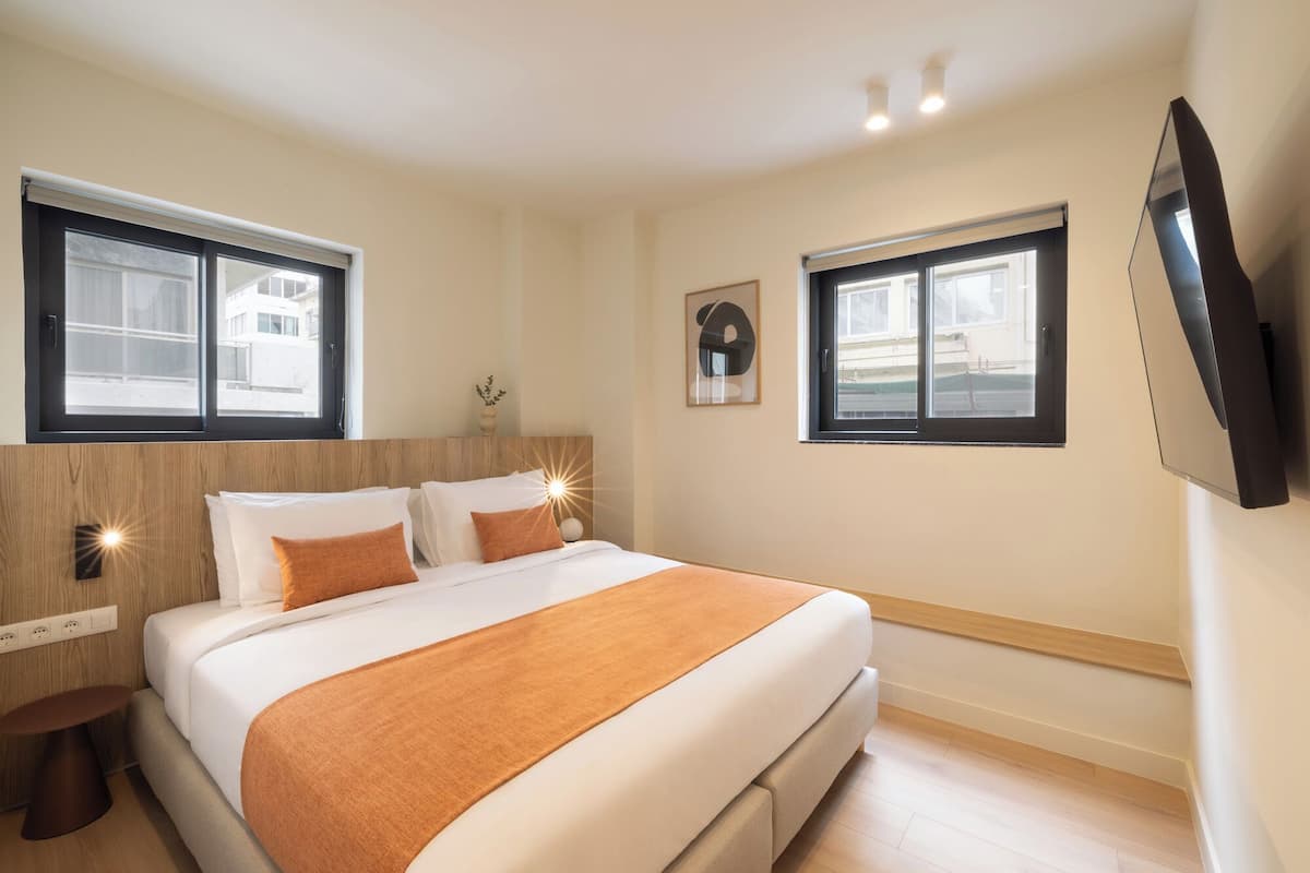 A modern hotel room with a large bed, two windows, a wall-mounted TV, and a bedside table.