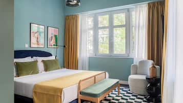 Ein Schlafzimmer mit einem Bett, einer Bank, einem Stuhl, einer Lampe und einem Fenster mit Vorhängen.