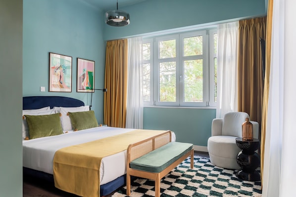 Ein Schlafzimmer mit einem Bett, einer Bank, einem Stuhl, einer Lampe und einem Fenster mit Vorhängen.