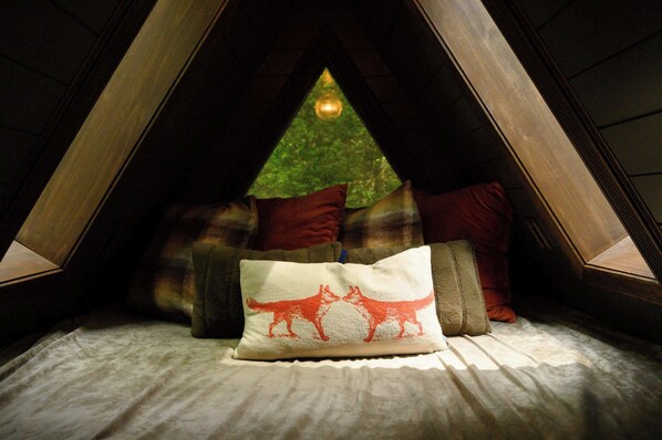 Een zolderkamer met een bed, kussens en uitzicht op een boom door een drielingsraam.