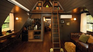 A loft-style room with a kitchen area, wooden flooring, a ladder, a couch, and a bookshelf. - Romantic AFrame| Firepit| Grill | Projector | Pets (Pocono Pines)