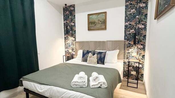 Un letto rifatto con cura, con copripiumino verde e due asciugamani piegati, una testiera in legno, un quadro incorniciato al muro e carta da parati con motivi floreali.