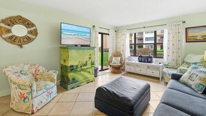 A living room with a beach-themed sofa, a flat-screen TV, and a view of the outdoors. - ✨Ocean and Pool Views | 1st Floor with Private Patio | Castle Reef 109✨ (New Smyrna Beach)