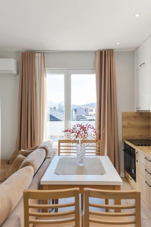 Un salon moderne comprenant un canapé, une table à manger et des chaises, un coin cuisine avec des placards et une cuisinière, ainsi que de grandes fenêtres avec des rideaux.