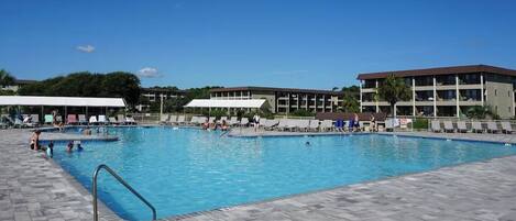 A large outdoor swimming pool with multiple lanes, surrounded by lounge chairs and umbrellas, and adjacent to a multi-story building.