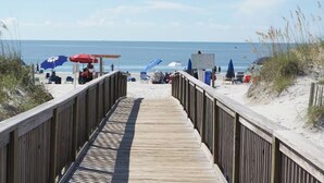 Ein hölzerner Steg führt zu einem Strand mit Sonnenschirmen und Liegestühlen.