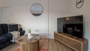Un salon moderne avec un canapé, une table basse, un téléviseur posé sur un meuble en bois, un miroir rond et un panneau décoratif en forme de sourire accroché au mur.