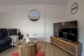 A modern living room with a sofa, a coffee table, a TV on a wooden stand, a round mirror, and a 'smile' sign on the wall.