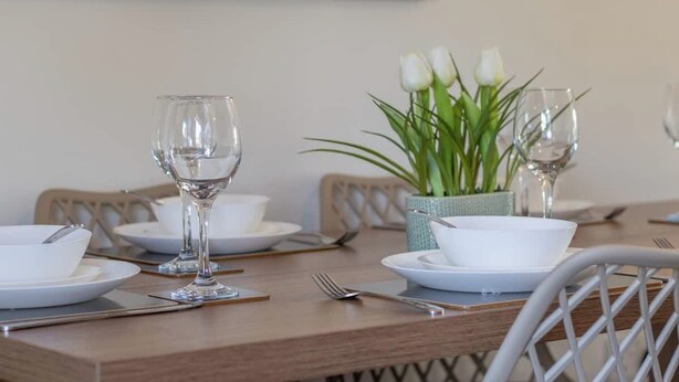 Une table à manger dressée avec de l’assiette blanche, des bols, des couverts et des verres à vin, et un vase rempli de fleurs blanches placé au centre.