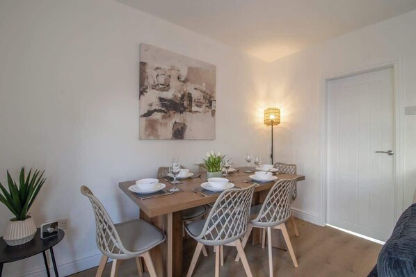 Un coin repas avec une table en bois dressée pour quatre personnes, entourée de chaises au dossier ajouré, d’une plante en pot et d’un tableau abstrait accroché au mur.