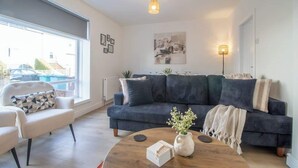 Un salon moderne avec un canapé bleu foncé, un fauteuil blanc, une table basse ronde en bois et une grande fenêtre donnant sur la rue.