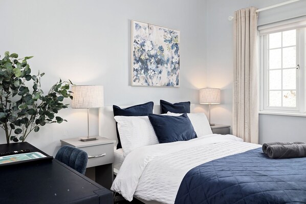 Une chambre à coucher avec un lit, des tables de chevet, des lampes, un tableau et une fenêtre avec des rideaux.