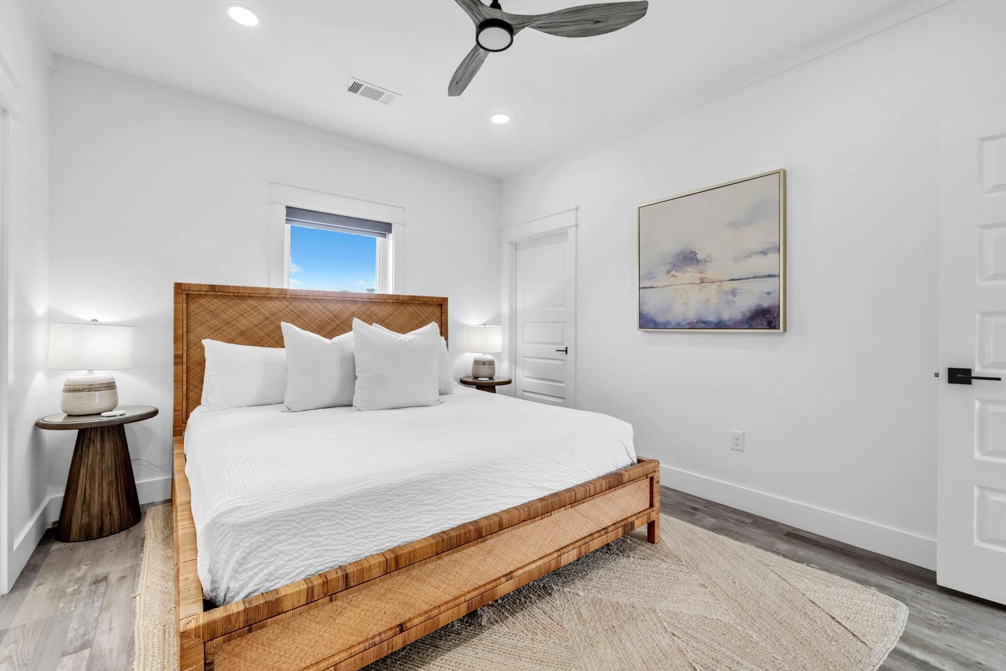 Un dormitorio con estructura de cama de madera, ropa de cama blanca, ventilador de techo, ventana y un cuadro en la pared.