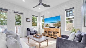 A spacious living room with a ceiling fan, a large TV, and a view of the beach through the windows. - Seapointe Serenity (Santa Rosa Beach)