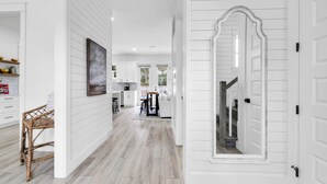 A modern kitchen with white cabinetry, a dining area with a wicker chair, and a hallway with a large mirror and white doors. - Seapointe Serenity (Santa Rosa Beach)