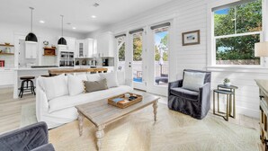 A spacious living area with a white sofa, a coffee table, and a grey armchair. There is a kitchen in the background with white cabinets and a dining area. - Seapointe Serenity (Santa Rosa Beach)