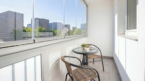 Ein Balkon mit rundem Tisch und zwei Korbstühlen, große Fenster mit Blick auf die Stadt und eine weiße Wand.
