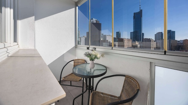 Ein Balkon mit Tisch und zwei Korbstühlen, einer Blumenvase und Blick auf die Stadt durch große Fenster.