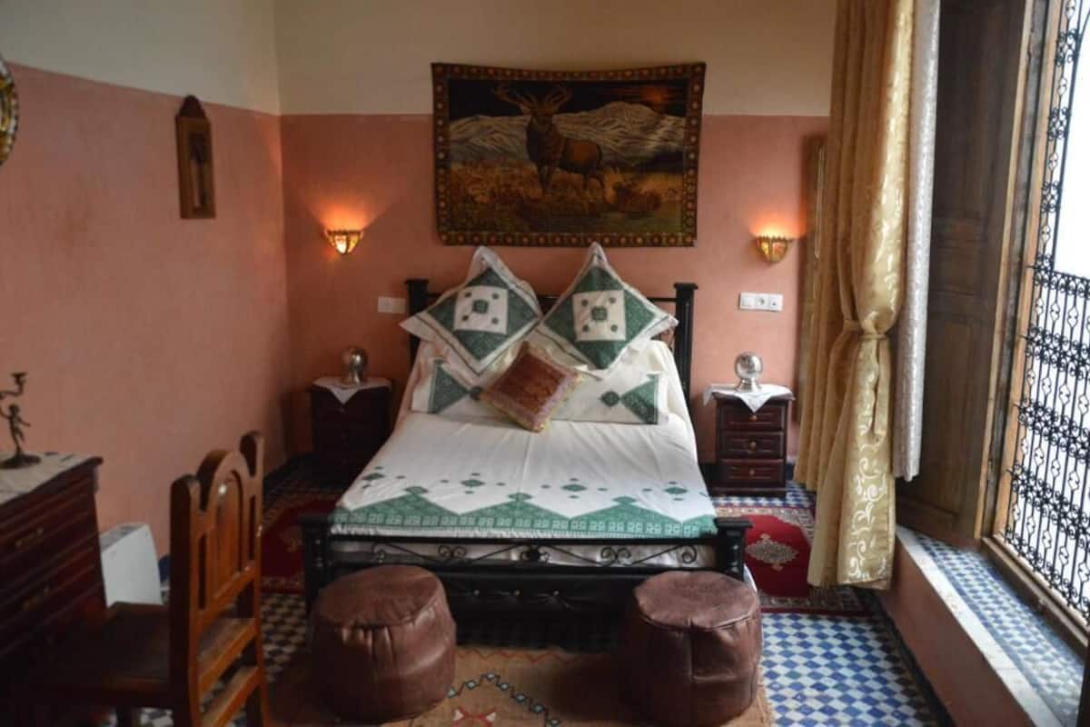 A bedroom with a bed, two nightstands, a wall-mounted lamp, a tapestry, and a window with a curtain.