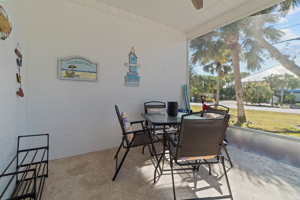 Ein überdachter Patio-Bereich mit Tisch und Stühlen, ein Schild mit der Aufschrift 'Our Beach House' und im Hintergrund eine Sicht auf Palmen und ein Gebäude.