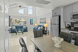 A modern kitchen with a refrigerator, microwave, and stove. There is a dining table with chairs and a decorative glass bowl. A blue sofa and a chair are in the living area with a window and artwork on the wall.