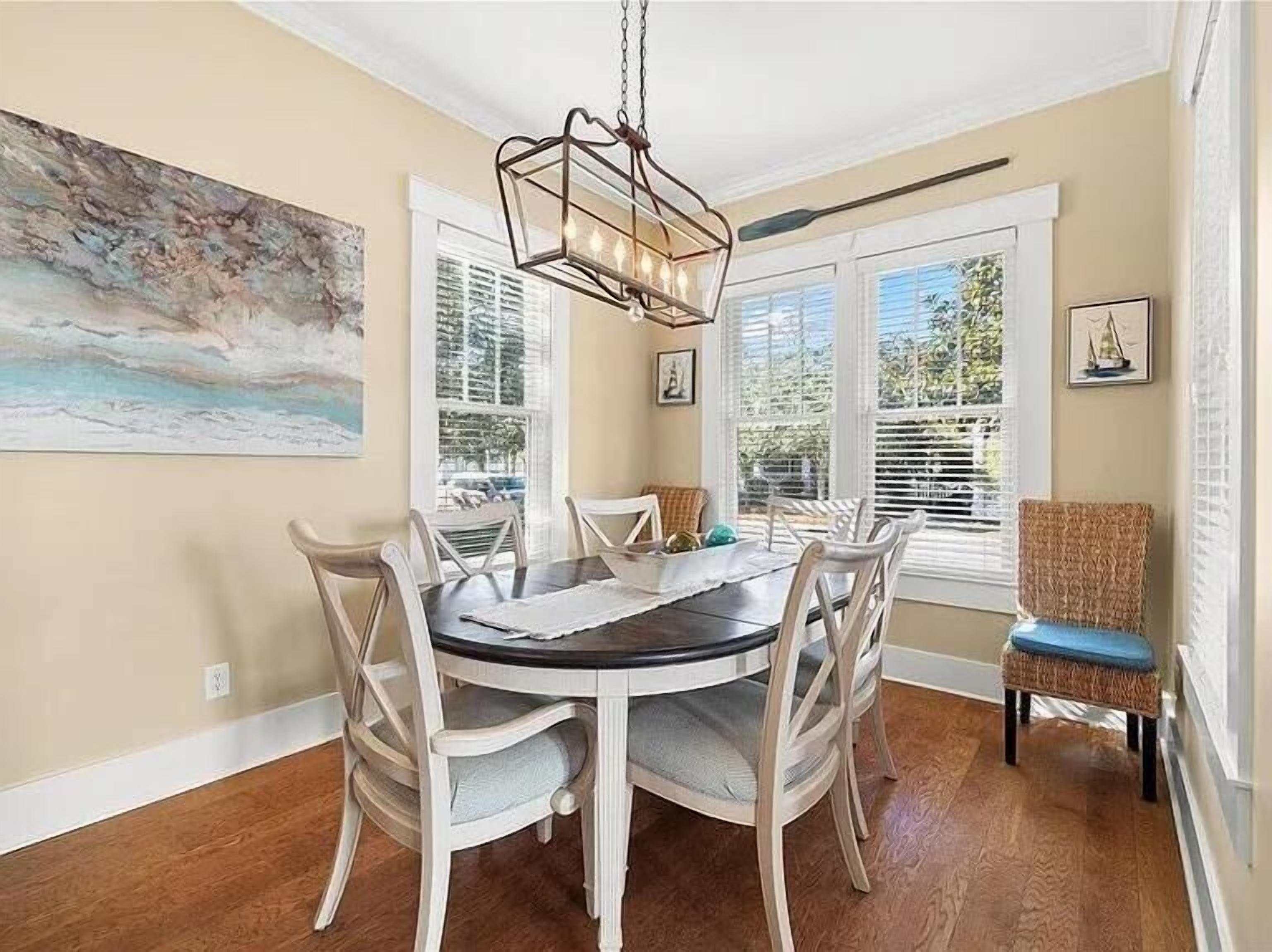 A dining area with a round table, six chairs, a hanging light fixture, and large windows with blinds.