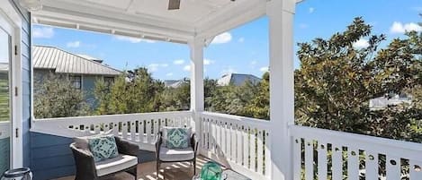 A covered porch with wicker furniture, a ceiling fan, and a view of surrounding buildings and trees.