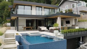 Ett modernt hus i två våningar med en swimmingpool, utomhusmöbler och en stor balkong med glasväggar.