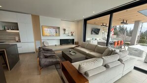 A modern living room with a sofa, coffee table, and a bench. There is a fireplace and a kitchen area visible in the background. - Above It All | Ocean Views + Spa Pool • Luxe 3BR/4.5BTH (North Vancouver)