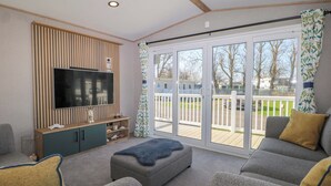 A living room with a flat-screen TV, grey sofas, a wooden entertainment unit, and a sliding glass door leading to an outdoor deck. - South Beach Landings 0028 (Rye)