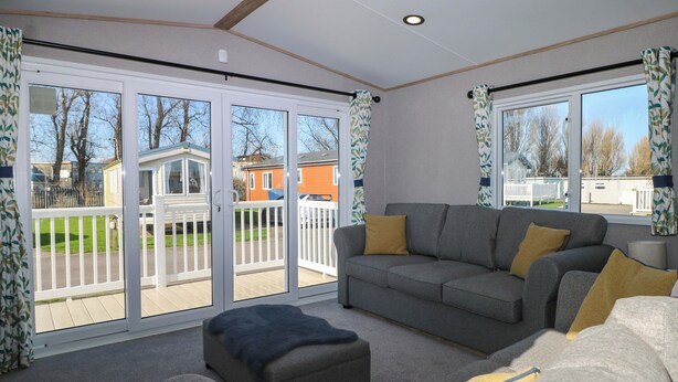 A living room with a grey sofa, a black ottoman, and a yellow throw pillow. There are large sliding glass doors leading to a balcony with a view of houses and a fence. - South Beach Landings 0028 (Rye)