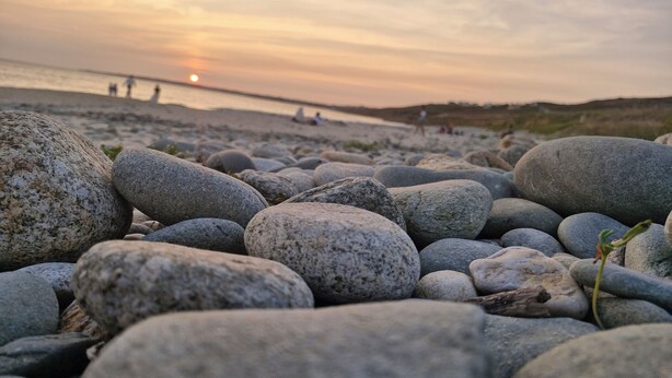 Une plage rocheuse, parsemée de gros galets, avec un coucher de soleil en arrière-plan.