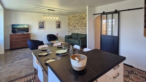 Une cuisine moderne dotée d’un îlot central, d’un coin repas avec des chaises et une table, d’un mur d’accent en pierre et d’un espace de vie avec un canapé et une télévision.